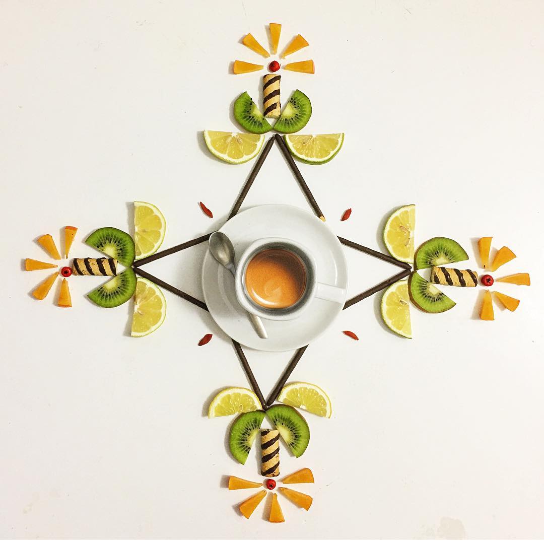 A whimsical arrangement of sliced fruits, chocolate, and a cup of espresso forming a symmetrical flower-like pattern