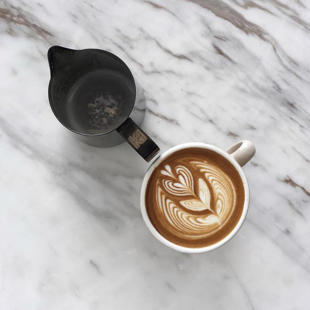 A freshly poured cup of cappuccino with intricate latte art next to a black pitcher on a marble surface