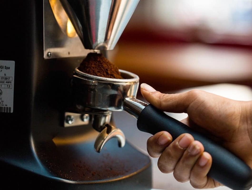 A barista's hand holds a perfectly dosed portafilter filled with fresh ground coffee