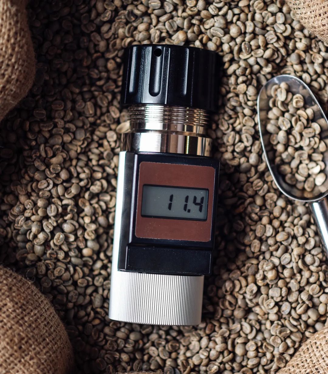 A coffee refractometer displaying a reading of 1.14 on its digital screen, surrounded by raw coffee beans.