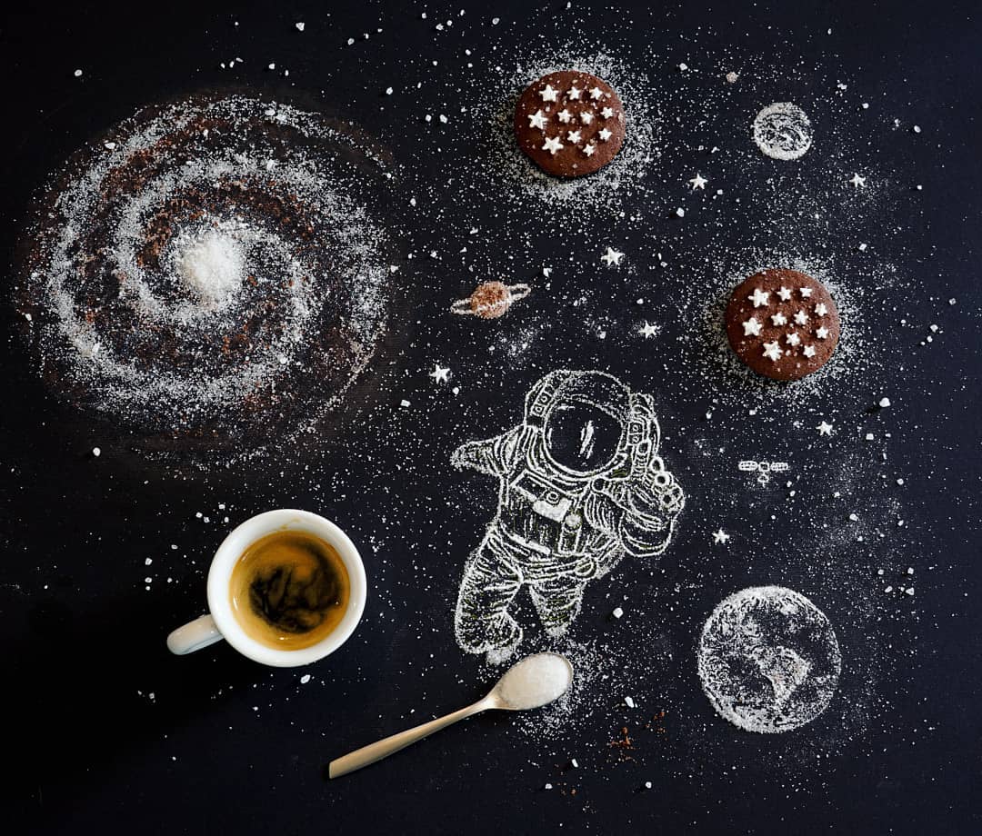 A whimsical culinary cosmos laid out on a dark surface, where powdered sugar planets orbit a coffee cup sun