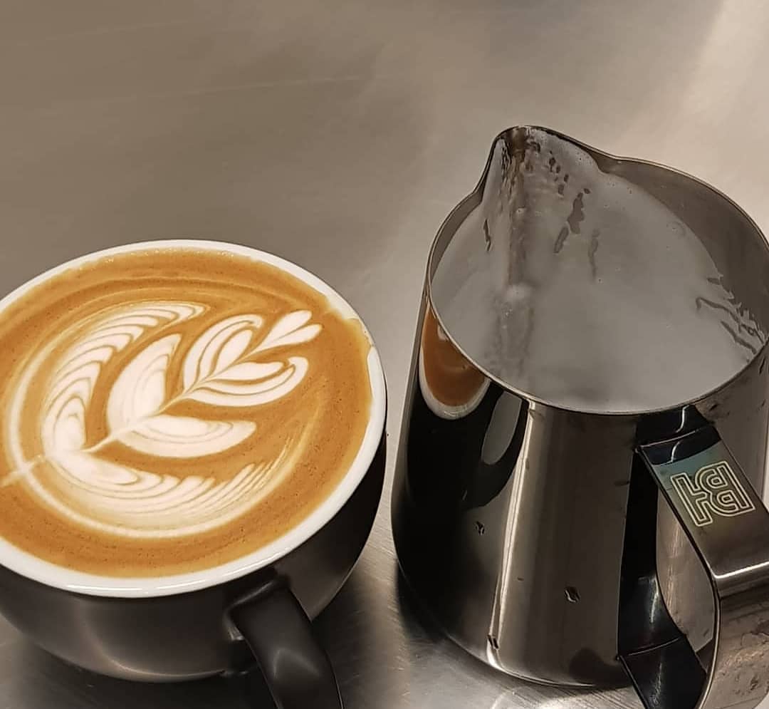 A freshly brewed cup of latte with artful milk foam design next to a metallic steaming pitcher