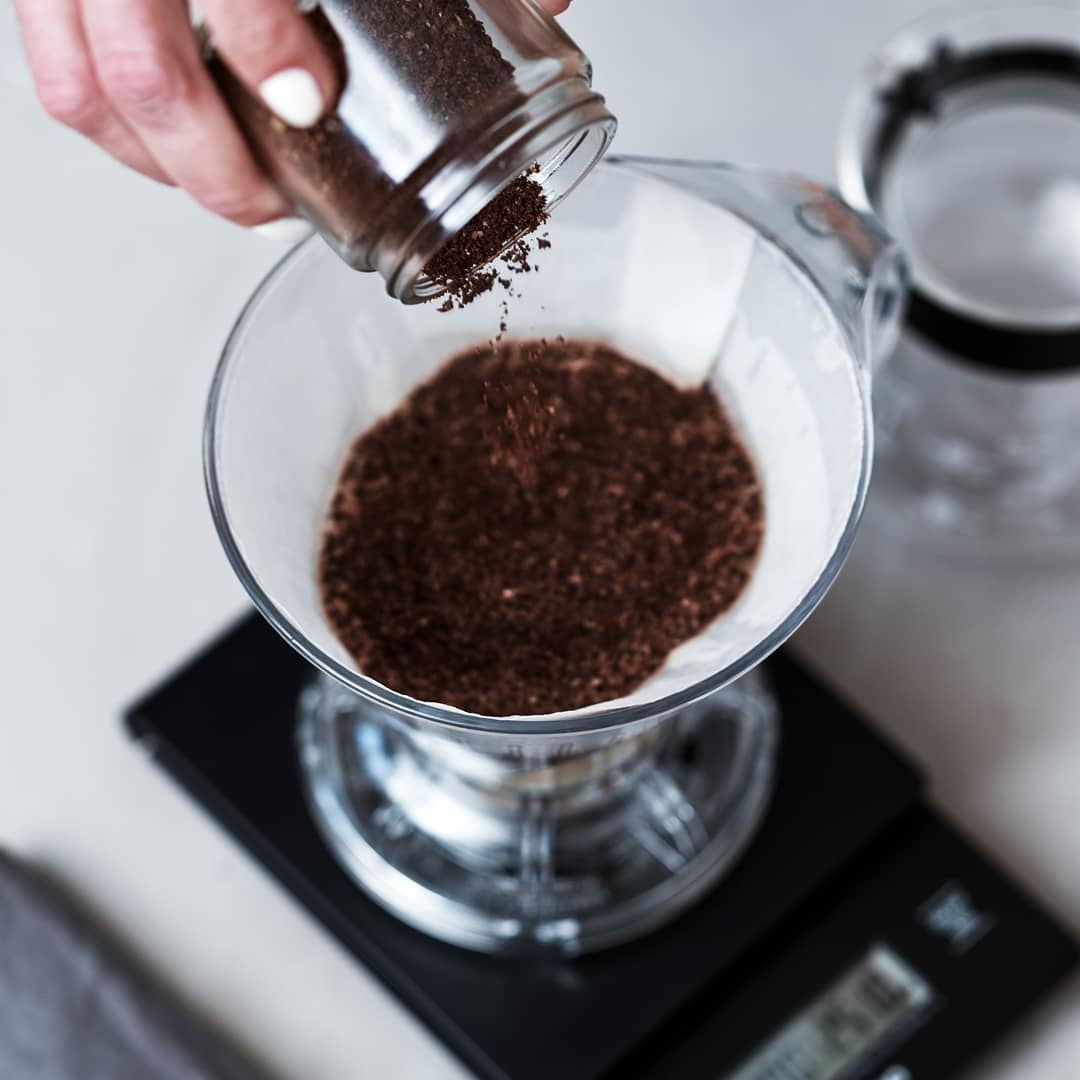 Person pouring ground coffee into a glass coffee dripper on a digital scale